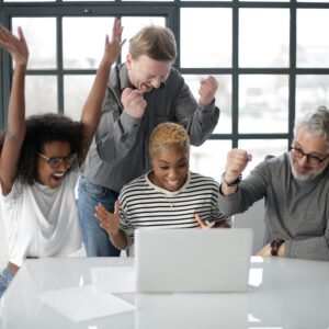 Cheerful multiethnic coworkers of different ages in casual clothing clenching fists and raising hands up in joy during successful teamwork in room